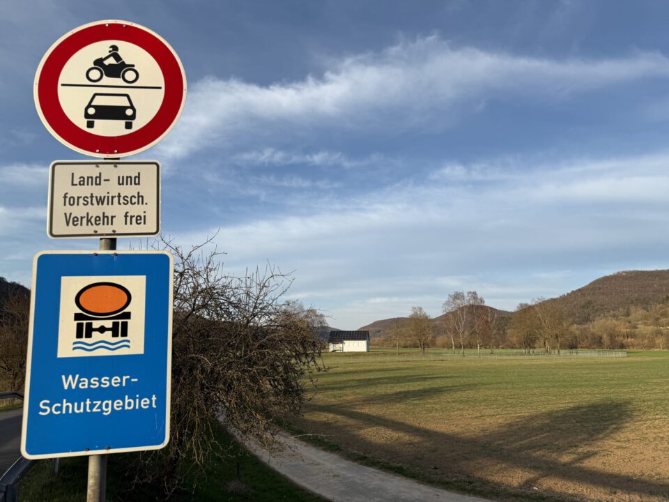 Das Bild zeigt eine ländliche Landschaft mit klarem Himmel und Hügeln im Hintergrund. Im Vordergrund stehen zwei Verkehrsschilder: eines verbietet die Durchfahrt für Motorräder und Autos, das andere weist auf ein Wasserschutzgebiet hin. Darunter befindet sich ein Zusatzschild, das land- und forstwirtschaftlichen Verkehr erlaubt.