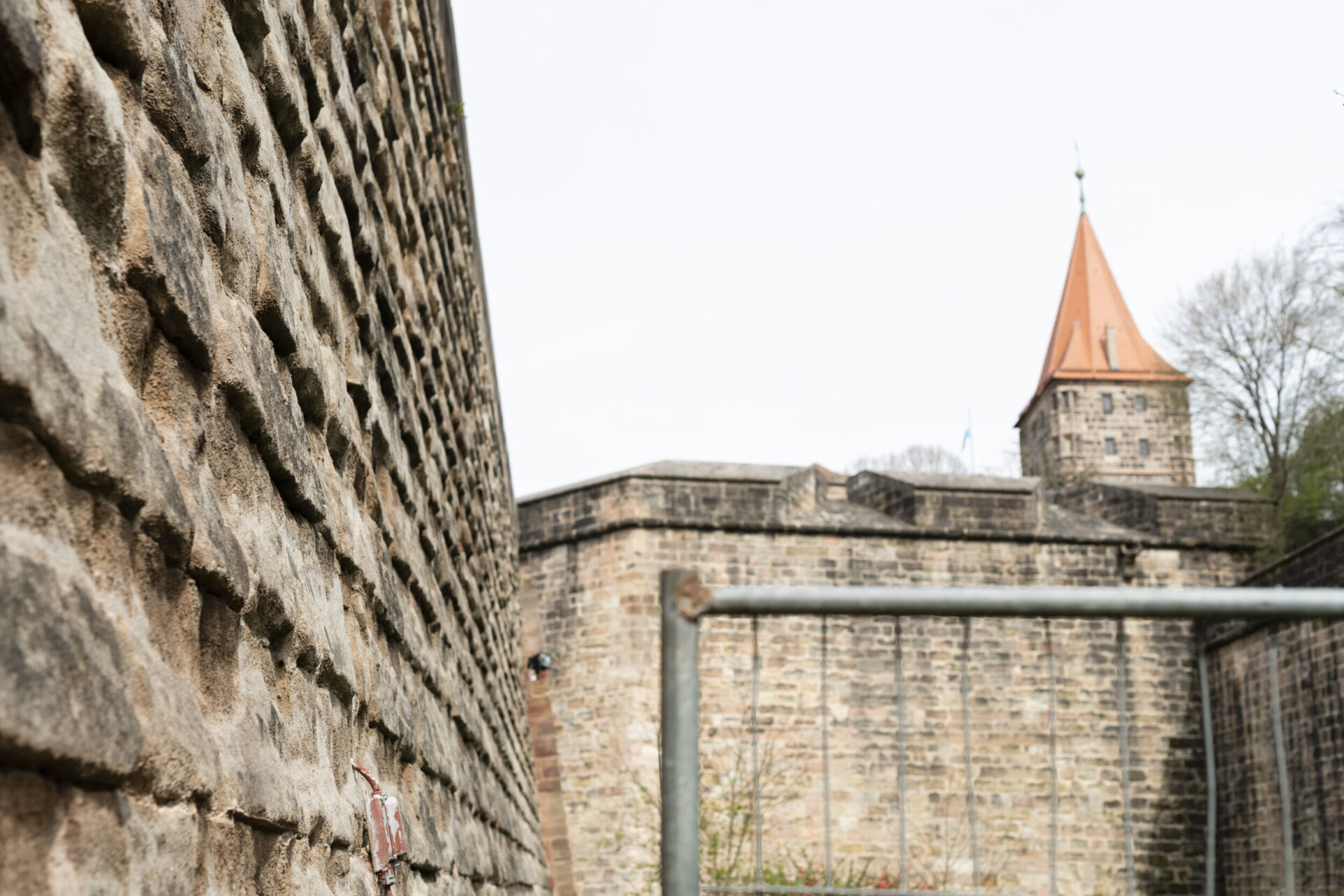 Das Bild zeigt eine Nahaufnahme einer Steinmauer auf der linken Seite, im Hintergrund sind eine weitere Mauer und ein Turm mit orangefarbenem Kegeldach zu sehen. In der Bildmitte befindet sich ein Metallgeländer oder -rahmen. Die Szene wirkt wie ein Teil einer alten Festungs- oder Schlossanlage.