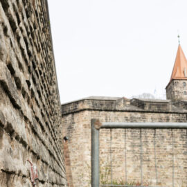 Das Bild zeigt eine Nahaufnahme einer Steinmauer auf der linken Seite, im Hintergrund sind eine weitere Mauer und ein Turm mit orangefarbenem Kegeldach zu sehen. In der Bildmitte befindet sich ein Metallgeländer oder -rahmen. Die Szene wirkt wie ein Teil einer alten Festungs- oder Schlossanlage.