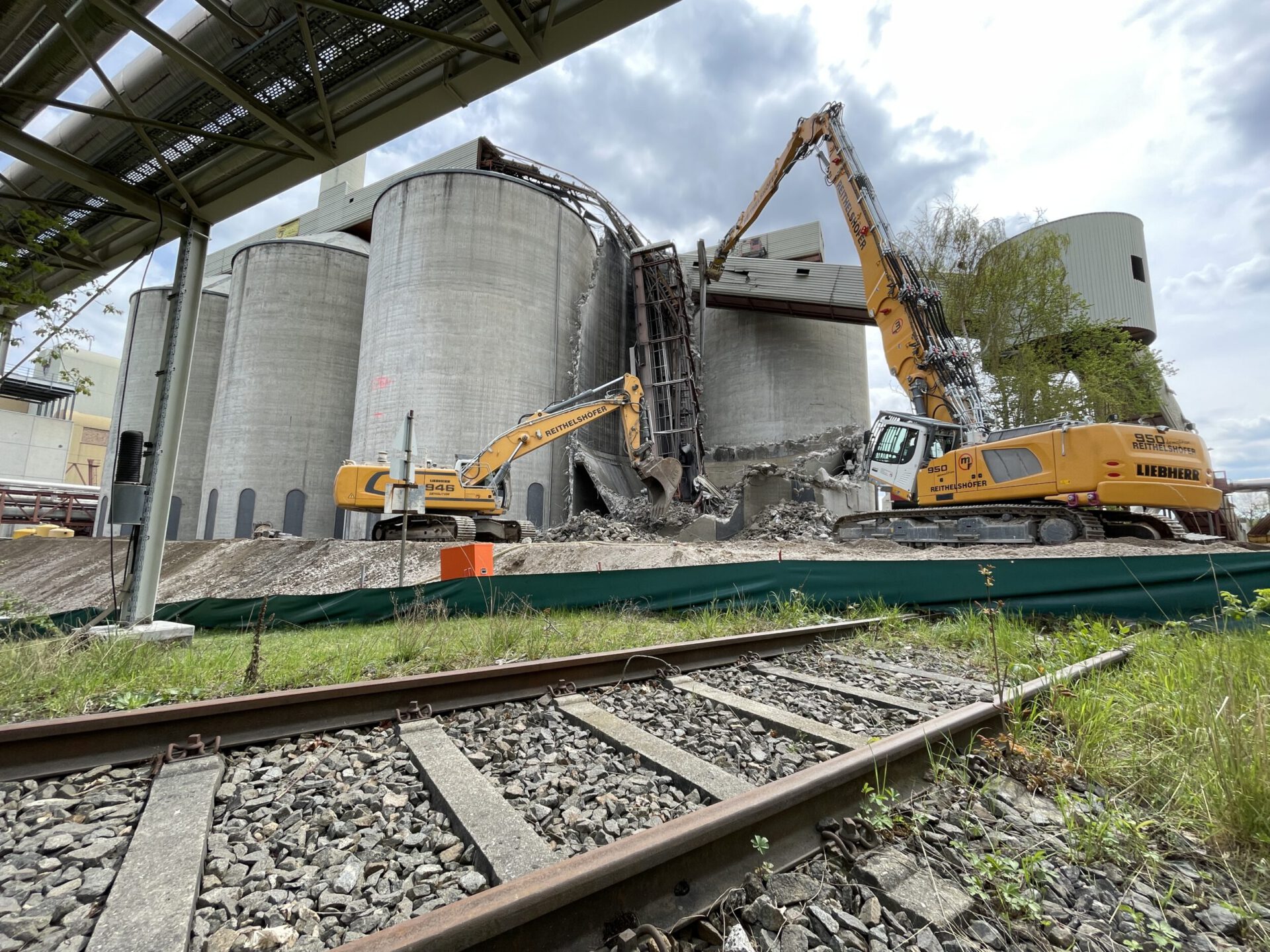 Auf dem Bild ist eine Baustelle zu sehen, auf der zwei große Bagger zylindrische Betonsilos abreißen. Im Vordergrund verläuft ein Gleis, und oben links ist eine Überbauung mit Rohrleitungen zu erkennen. Die Szene spielt im Freien unter einem bewölkten Himmel.