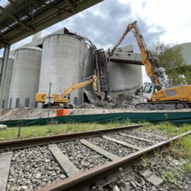 Auf dem Bild ist eine Baustelle zu sehen, auf der zwei große Bagger zylindrische Betonsilos abreißen. Im Vordergrund verläuft ein Gleis, und oben links ist eine Überbauung mit Rohrleitungen zu erkennen. Die Szene spielt im Freien unter einem bewölkten Himmel.