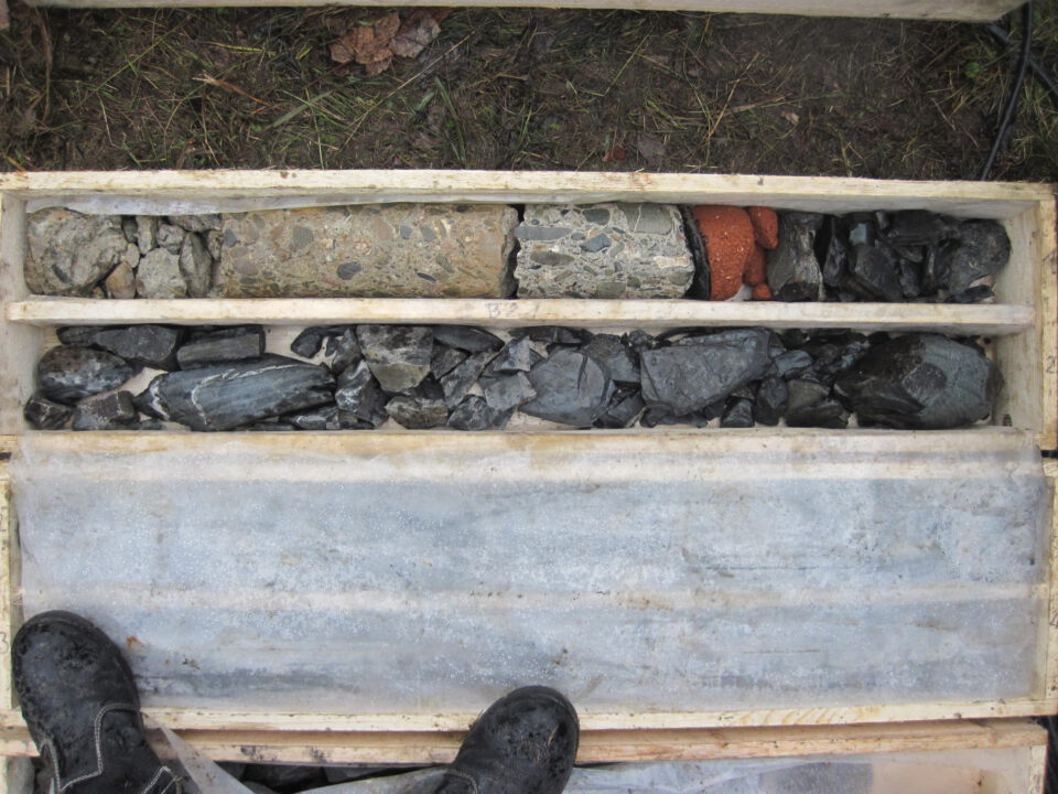 Das Bild zeigt eine Holzkiste mit drei Fächern, in denen verschiedene Gesteinsproben aufbewahrt werden. Im oberen Fach liegt ein zylindrischer Bohrkern mit einer Mischung aus Beton, Gestein und rötlichem Material, das mittlere Fach enthält unterschiedlich große dunkelgraue Steine. Das untere Fach ist leer oder mit einer Schutzschicht abgedeckt, am unteren Bildrand sind zwei Arbeitsschuhe zu sehen.