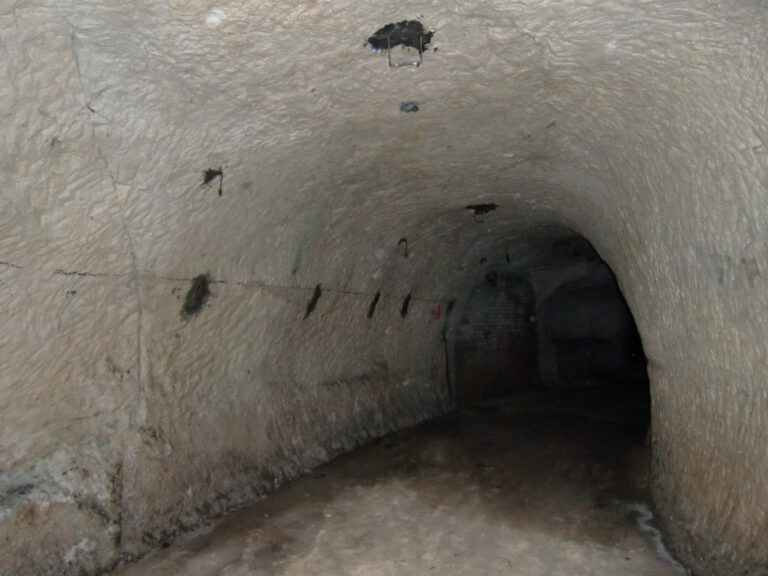 Das Bild zeigt einen unterirdischen Tunnel mit gewölbter Decke und unebenen, feuchten Boden. Die Wände und die Decke bestehen aus grobem Stein oder Beton und weisen mehrere kleine Öffnungen oder Vertiefungen auf.