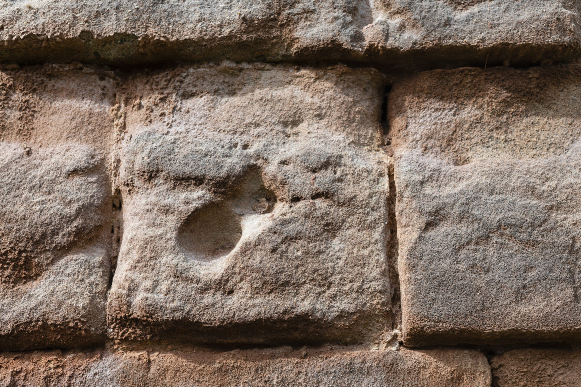 Nahaufnahme einer Steinmauer aus rechteckigen Blöcken mit rauer, verwitterter Oberfläche. Einer der Steine weist eine deutliche Vertiefung oder ein Loch auf.