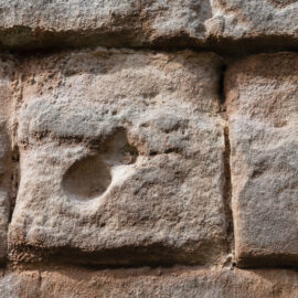 Nahaufnahme einer Steinmauer aus rechteckigen Blöcken mit rauer, verwitterter Oberfläche. Einer der Steine weist eine deutliche Vertiefung oder ein Loch auf.
