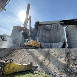 Auf dem Bild ist eine Baustelle mit großen, zylindrischen Betonbauten zu sehen. Ein gelber Bagger mit Hydraulikhammer bearbeitet eines der Bauwerke, das bereits teilweise abgetragen ist. Im Vordergrund liegen weitere Baumaschinenteile auf einem Schotterhaufen, während die Sonne hell über der Szene scheint.