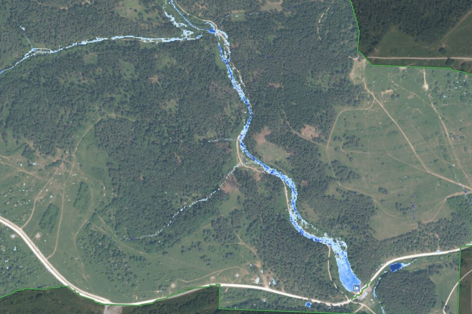 Das Bild zeigt eine Luftaufnahme einer Landschaft mit einem sich schlängelnden Fluss, der in Blau hervorgehoben ist und von der Bildmitte oben nach rechts unten verläuft. Die Umgebung besteht aus dichten Wäldern, offenen Feldern und vereinzelt stehenden Gebäuden. Straßen sind ebenfalls sichtbar, darunter eine markante Straße im unteren Bereich des Bildes und eine weitere, die diese im rechten Bereich kreuzt.