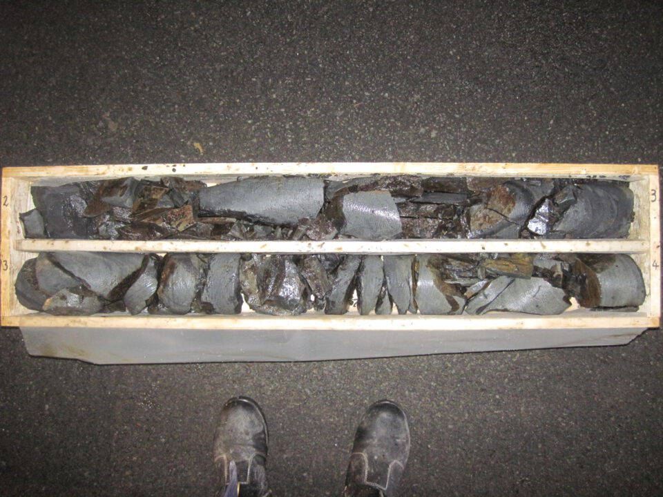 Das Bild zeigt eine Holzkiste mit zwei Reihen von Bohrkernproben aus Gestein. Die Proben sind dunkelgrau, teilweise glänzend und in Segmente unterteilt. Die Kiste ist seitlich mit den Zahlen „2“ und „3“ sowie „3“ und „4“ beschriftet, am unteren Bildrand sind zwei Arbeitsschuhe zu sehen.