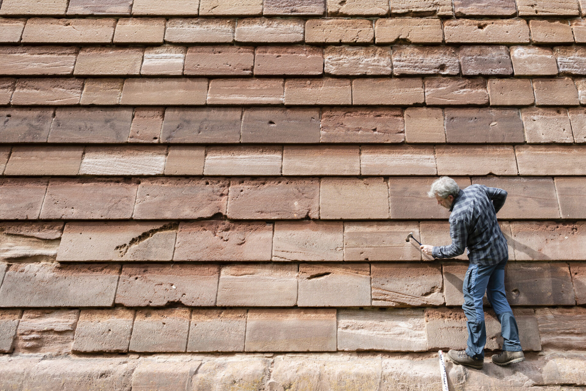 Eine Person arbeitet an einer großen Wand aus rechteckigen Steinplatten in verschiedenen Braun- und Beigetönen. Die Person hält einen Hammer und scheint die Platten zu überprüfen oder zu reparieren. Einige der Steine zeigen sichtbare Abnutzungs- oder Schadensspuren.