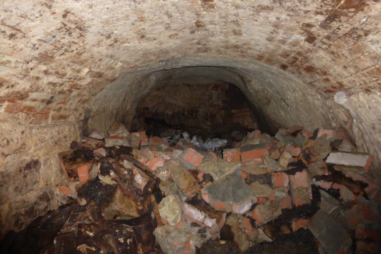 Das Bild zeigt einen unterirdischen Tunnel oder Keller mit einer gewölbten Decke aus Ziegelsteinen. Auf dem Boden liegt ein großer Haufen aus Schutt, bestehend aus zerbrochenen Ziegeln, Steinen und weiteren Trümmern.