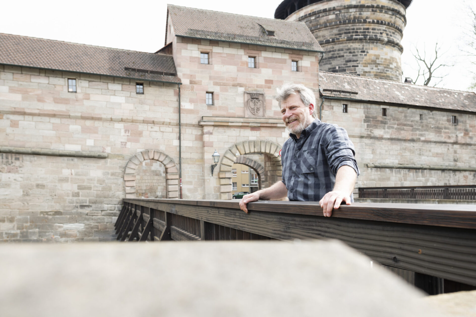 Eine Person steht auf einer Holzbrücke vor einem alten Steingebäude mit gewölbten Türen und Fenstern. Im Hintergrund ist ein rundes Turmgebäude mit Ziegeldach zu sehen. Die Person trägt ein kariertes Hemd, das Gesicht ist unscharf.