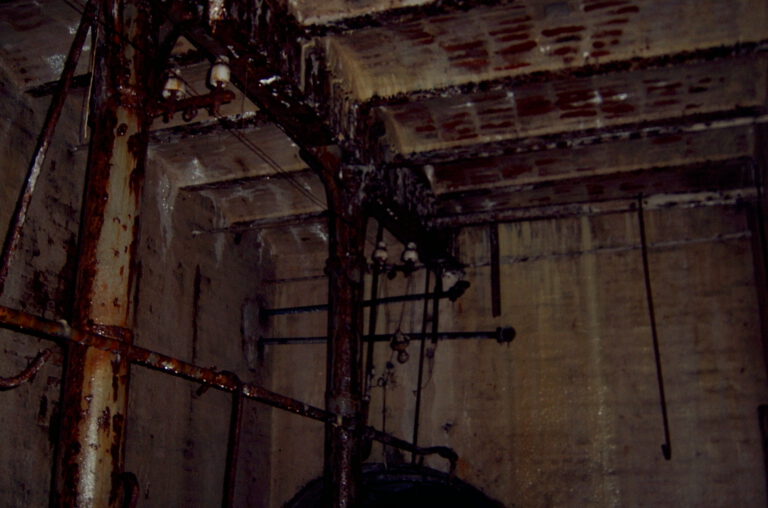 Das Bild zeigt einen alten, verrosteten Keller- oder Maschinenraum mit Metallrohren und Trägern. Wände und Decke bestehen aus Beton und weisen deutliche Wasserschäden und Rostflecken auf. Verschiedene Rohre, Ketten und mechanische Bauteile hängen von der Decke oder sind an den Wänden befestigt.