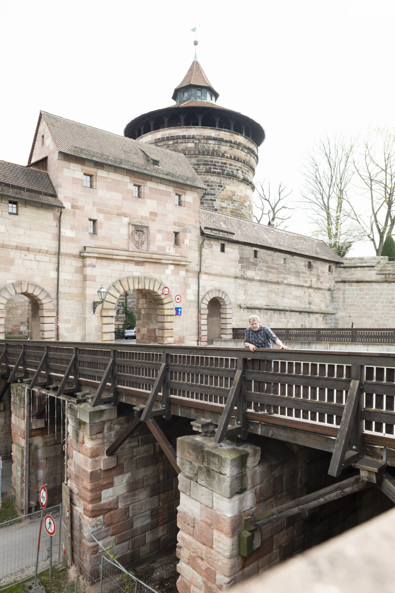 Das Bild zeigt ein historisches Steingebäude mit einem Turm, vermutlich Teil einer Burg oder Befestigungsanlage. Eine Holzbrücke führt zu gewölbten Eingängen, an denen Verkehrsschilder für Fußgänger und Fahrzeuge angebracht sind. Eine Person lehnt am Brückengeländer und blickt nach unten, im Hintergrund stehen laublose Bäume.