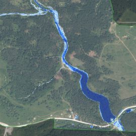 Das Bild zeigt eine Luftaufnahme einer Landschaft mit einem sich schlängelnden Fluss, der in Blau hervorgehoben ist und von der Bildmitte oben nach rechts unten verläuft. Die Umgebung besteht aus dichten Wäldern, offenen Feldern und vereinzelt stehenden Gebäuden. Straßen sind ebenfalls sichtbar, darunter eine markante Straße im unteren Bereich des Bildes und eine weitere, die diese im rechten Bereich kreuzt.