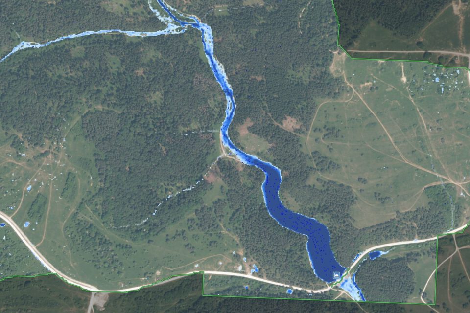 Das Bild zeigt eine Luftaufnahme einer Landschaft mit einem sich schlängelnden Fluss, der in Blau hervorgehoben ist und von der Bildmitte oben nach rechts unten verläuft. Die Umgebung besteht aus dichten Wäldern, offenen Feldern und vereinzelt stehenden Gebäuden. Straßen sind ebenfalls sichtbar, darunter eine markante Straße im unteren Bereich des Bildes und eine weitere, die diese im rechten Bereich kreuzt.