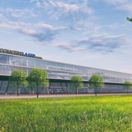 Das Bild zeigt ein modernes, mehrstöckiges Gebäude mit großen Glasfenstern und einer klaren, geradlinigen Architektur. Vor dem Gebäude erstreckt sich eine gepflegte Rasenfläche mit mehreren Bäumen. Der Himmel ist teils bewölkt mit sichtbaren blauen Abschnitten.