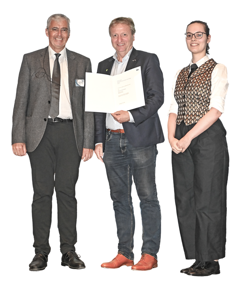 Drei Personen stehen nebeneinander. Die mittlere Person hält ein teilweise sichtbares Dokument oder eine Urkunde in den Händen. Alle drei tragen unterschiedliche formelle bis legere Kleidung.