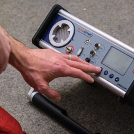 Das Bild zeigt eine Hand, die ein Gerät mit der Aufschrift „AquaGUARD“ über ein Bedienfeld steuert. Das Gerät verfügt über verschiedene Tasten, Drehregler, Anschlüsse sowie einen Bildschirm und liegt auf einem Teppichboden.