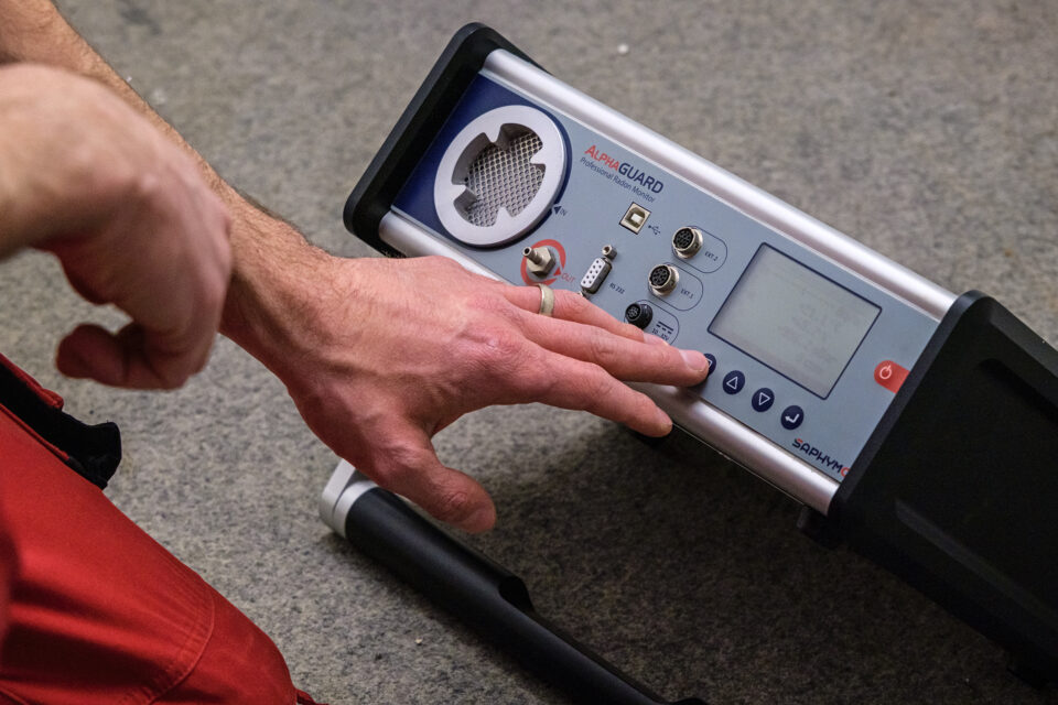 Das Bild zeigt eine Hand, die ein Gerät mit der Aufschrift „AquaGUARD“ über ein Bedienfeld steuert. Das Gerät verfügt über verschiedene Tasten, Drehregler, Anschlüsse sowie einen Bildschirm und liegt auf einem Teppichboden.