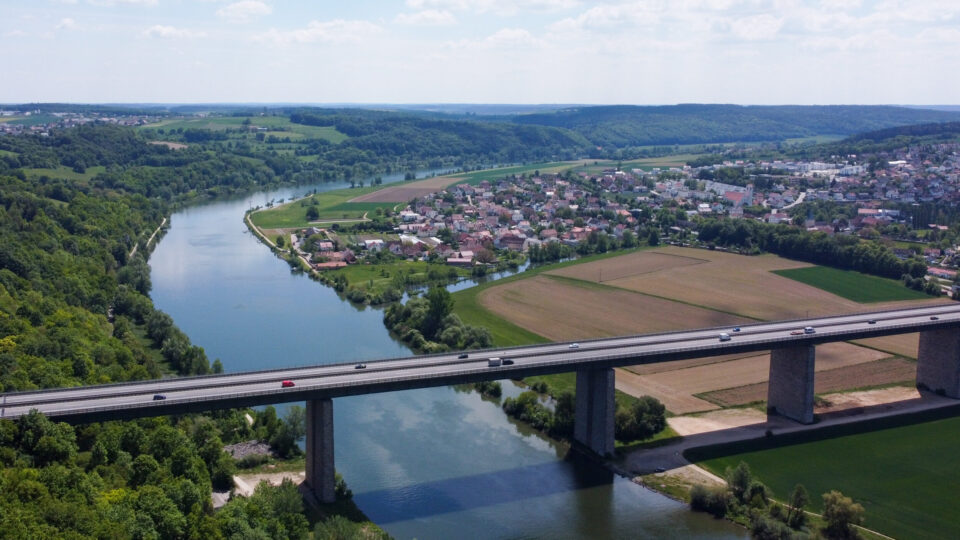 Das Bild zeigt eine Luftaufnahme eines Flusses, der sich durch eine Landschaft mit grünen Hügeln und Feldern schlängelt. Eine Brücke überquert den Fluss, auf der mehrere Fahrzeuge unterwegs sind. Auf einer Seite des Flusses liegt eine Stadt mit vielen Häusern und Gebäuden, umgeben von landwirtschaftlichen Flächen und Waldstücken.