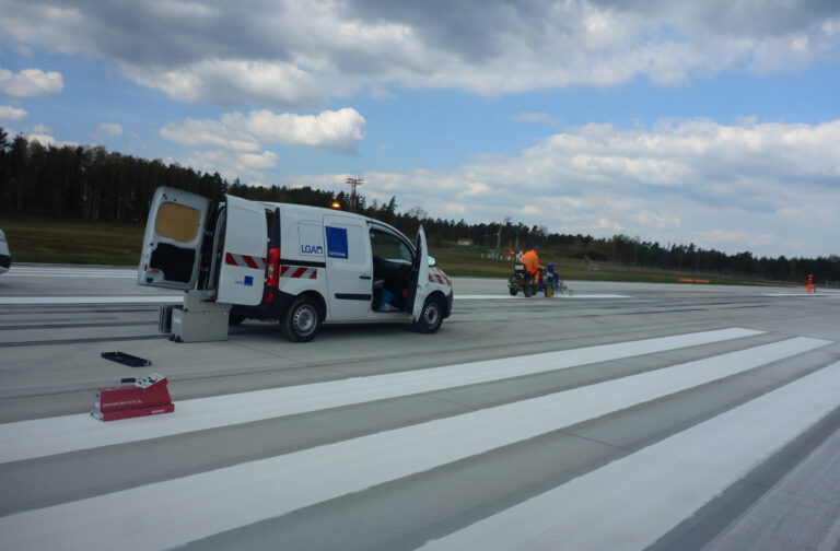 Das Bild zeigt eine Szene auf einer großen asphaltierten Fläche, vermutlich einer Start- oder Landebahn. Ein weißer Transporter mit geöffnetem Heck steht am Rand, während eine Person in orangefarbener Jacke weiter entfernt an einer Maschine arbeitet. Im Hintergrund sind Bäume und ein teils bewölkter Himmel zu sehen.