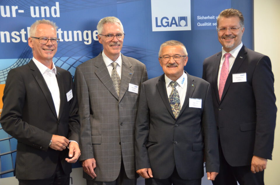 Vier Männer in formeller Geschäftskleidung stehen vor einem blauen Hintergrund mit Text und Logos. Sie tragen Anzüge, Krawatten und Namensschilder. Der Hintergrund zeigt unter anderem ein Logo und einen Slogan.