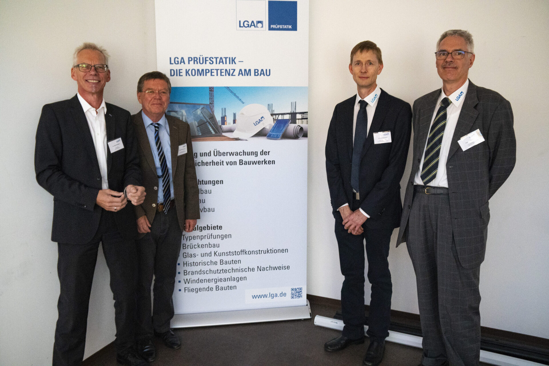 Vier Männer stehen vor einem Banner, das Informationen zu Leistungen und Tätigkeitsgebieten im Bereich der Bauwerksprüfung zeigt. Der Hintergrund enthält eine Auflistung verschiedener Bauarten und Prüfbereiche. Die Männer tragen formelle Kleidung.