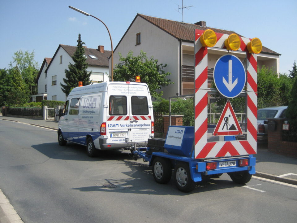 Das Bild zeigt einen weißen Transporter mit einem blauen Anhänger, der auf einer Wohnstraße geparkt ist. Auf dem Anhänger ist ein großes Verkehrsschild mit gelbem Blinklicht, einem blauen Richtungspfeil und einem Baustellen-Warnzeichen montiert. Im Hintergrund sind Wohnhäuser und Grünflächen zu sehen.
