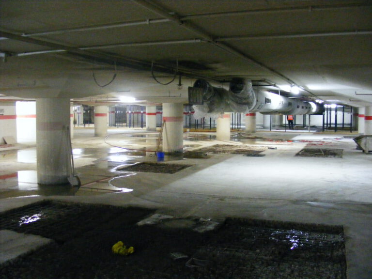 Das Bild zeigt eine Tiefgarage oder einen Kellerbereich, der sich im Bau oder in der Sanierung befindet. Mehrere Betonsäulen stützen die Decke, an der große Lüftungskanäle verlaufen. Auf dem teilweise nassen Boden sind Pfützen, freigelegter Untergrund sowie Baugeräte und Materialien verteilt.