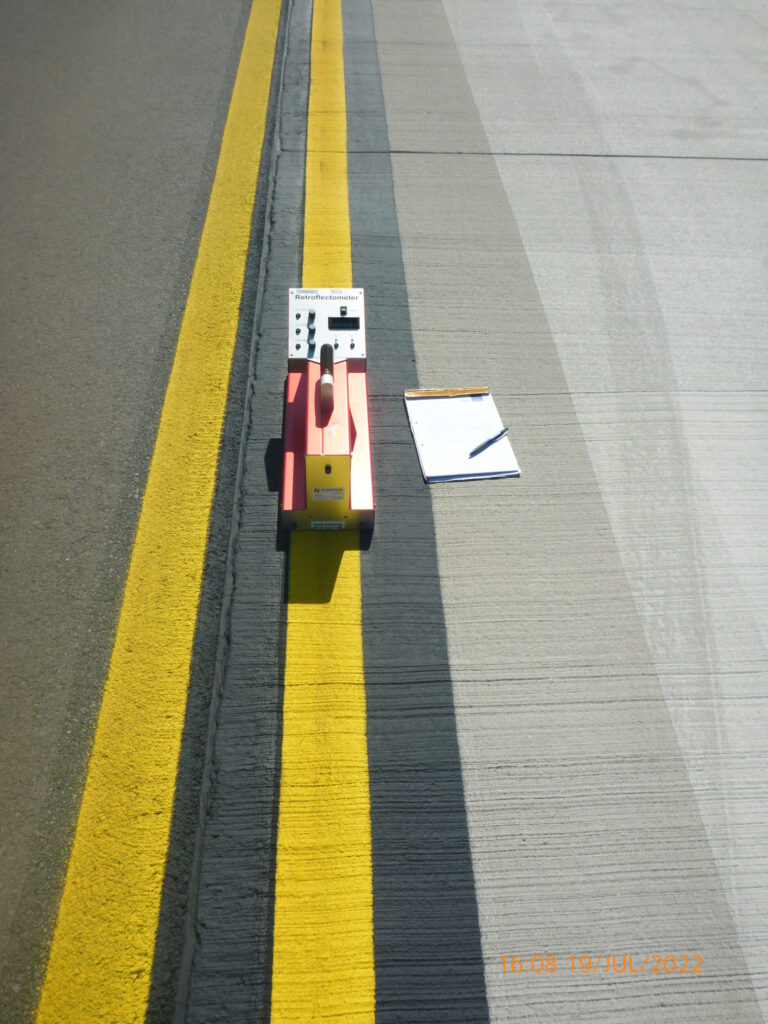 Das Bild zeigt einen Straßenabschnitt mit einer gelben Linie am Rand. Auf der Linie steht ein orangefarbenes Messgerät, daneben liegen ein Klemmbrett mit Papier und ein Stift.