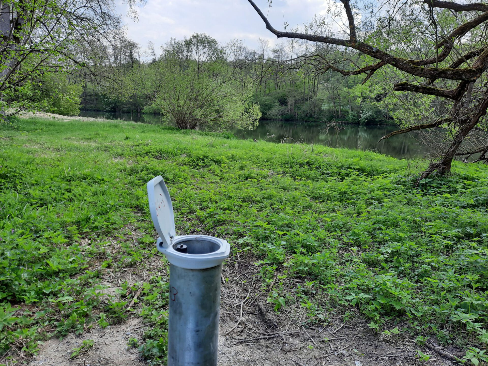 Das Bild zeigt eine grüne Wiesenfläche mit Bäumen und einem Gewässer im Hintergrund. Im Vordergrund steht ein zylindrischer Metallbehälter mit offenem Deckel, vermutlich ein Außenbehälter oder Abfallbehälter. Die Umgebung ist von dichter Vegetation geprägt und wirkt naturnah und ruhig.
