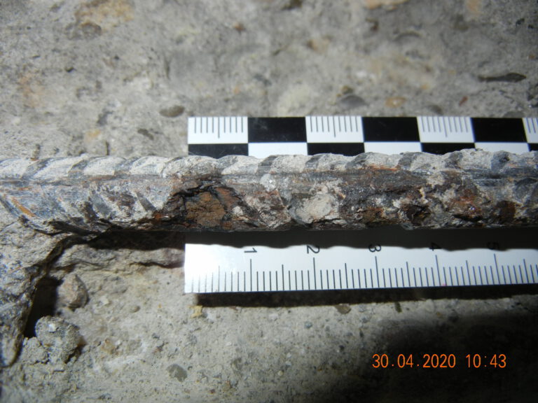 Das Bild zeigt eine Nahaufnahme eines korrodierten Bewehrungsstabs, der in Beton eingebettet ist. Zwei Messskalen liegen zur Referenz daneben – eine schwarz-weiße mit abwechselnden Quadraten und eine weiße mit Millimetereinteilung.