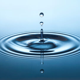 Das Bild zeigt eine Nahaufnahme eines Wassertropfens, der in eine Wasseroberfläche fällt und dabei konzentrische Wellen erzeugt. Der Tropfen ist in der Luft eingefangen, kurz bevor er das Wasser berührt. Der Hintergrund verläuft in einem sanften Farbverlauf von Hell- zu Dunkelblau.