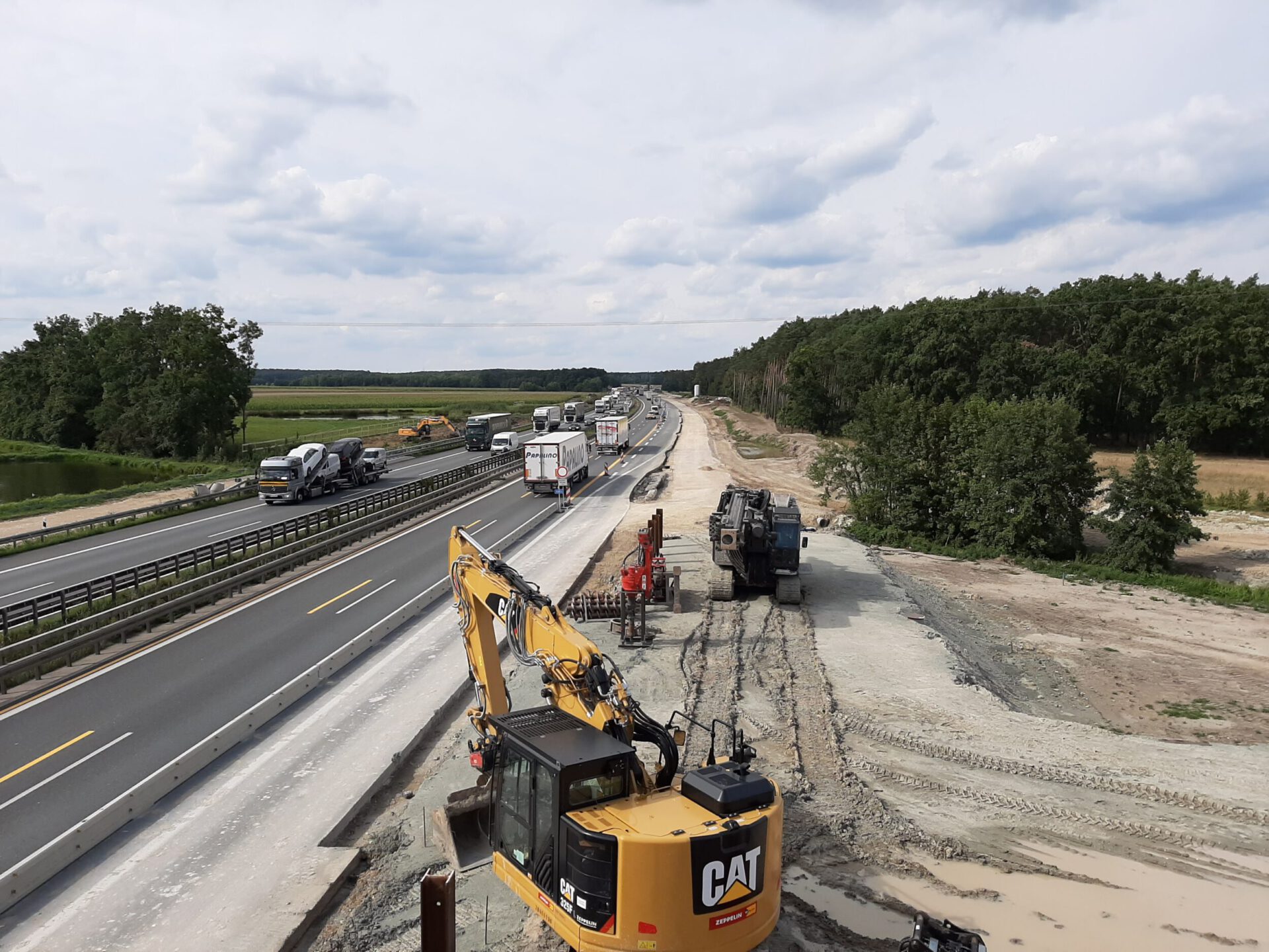 Das Bild zeigt eine Baustelle an einer Autobahn mit zwei Fahrspuren auf der linken Seite, auf denen mehrere Fahrzeuge wie Lastwagen und Autos unterwegs sind. Rechts davon befindet sich der Baustellenbereich mit Baufahrzeugen wie Baggern und weiterer Ausrüstung. Die Umgebung besteht aus Bäumen und offenen Feldern unter einem teilweise bewölkten Himmel.