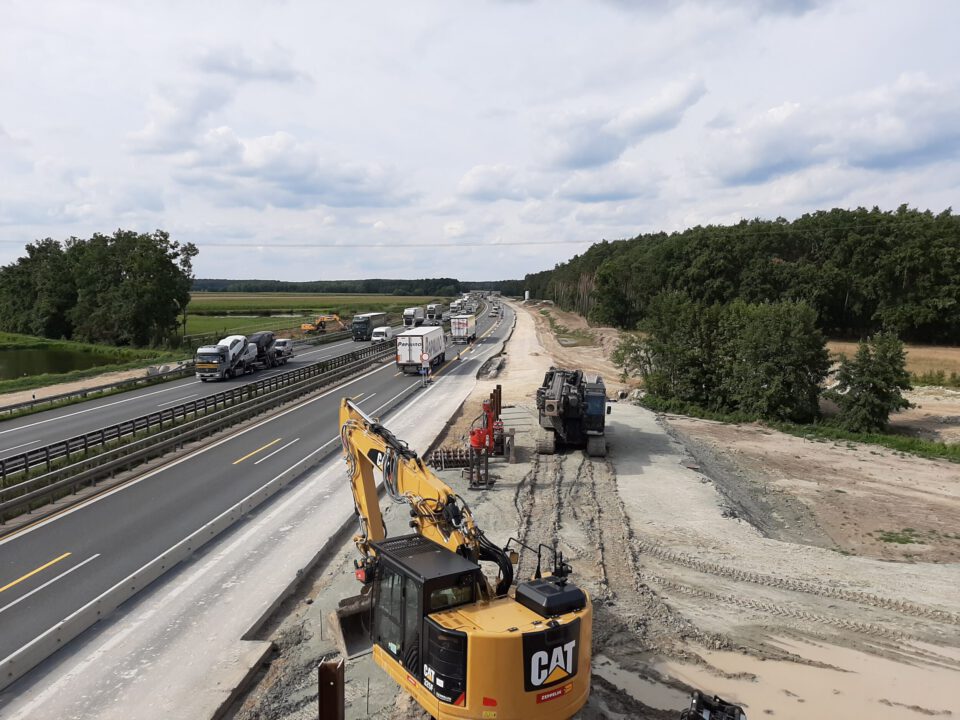 Das Bild zeigt eine Baustelle an einer Autobahn mit zwei Fahrspuren auf der linken Seite, auf denen mehrere Fahrzeuge wie Lastwagen und Autos unterwegs sind. Rechts davon befindet sich der Baustellenbereich mit Baufahrzeugen wie Baggern und weiterer Ausrüstung. Die Umgebung besteht aus Bäumen und offenen Feldern unter einem teilweise bewölkten Himmel.