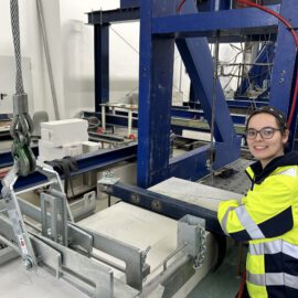 Das Bild zeigt eine industrielle Umgebung mit einer Person in Warnkleidung neben einer großen blauen Metallkonstruktion. Die Konstruktion gehört zu einer mechanischen oder fertigungstechnischen Anlage und enthält verschiedene Kabel, Haken und Maschinenteile.