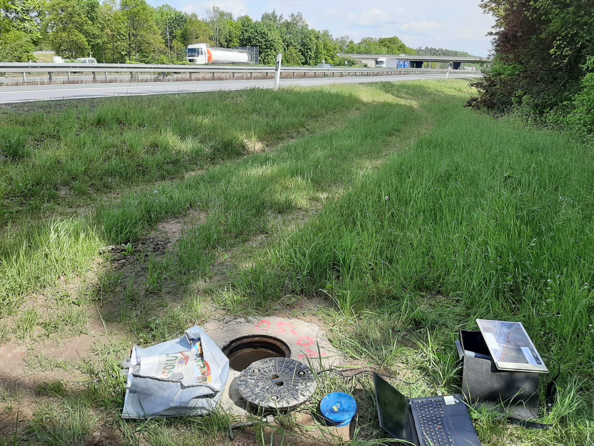 Das Bild zeigt eine grasbewachsene Fläche neben einer Autobahn, auf der Fahrzeuge vorbeifahren. Im Vordergrund befindet sich ein offener Kanalschacht mit dem Deckel daneben sowie verschiedene Gegenstände wie ein Laptop, ein blaues Messgerät und eine Tasche. Die Szene deutet auf eine Wartungs- oder Messaktivität im Außeneinsatz hin.