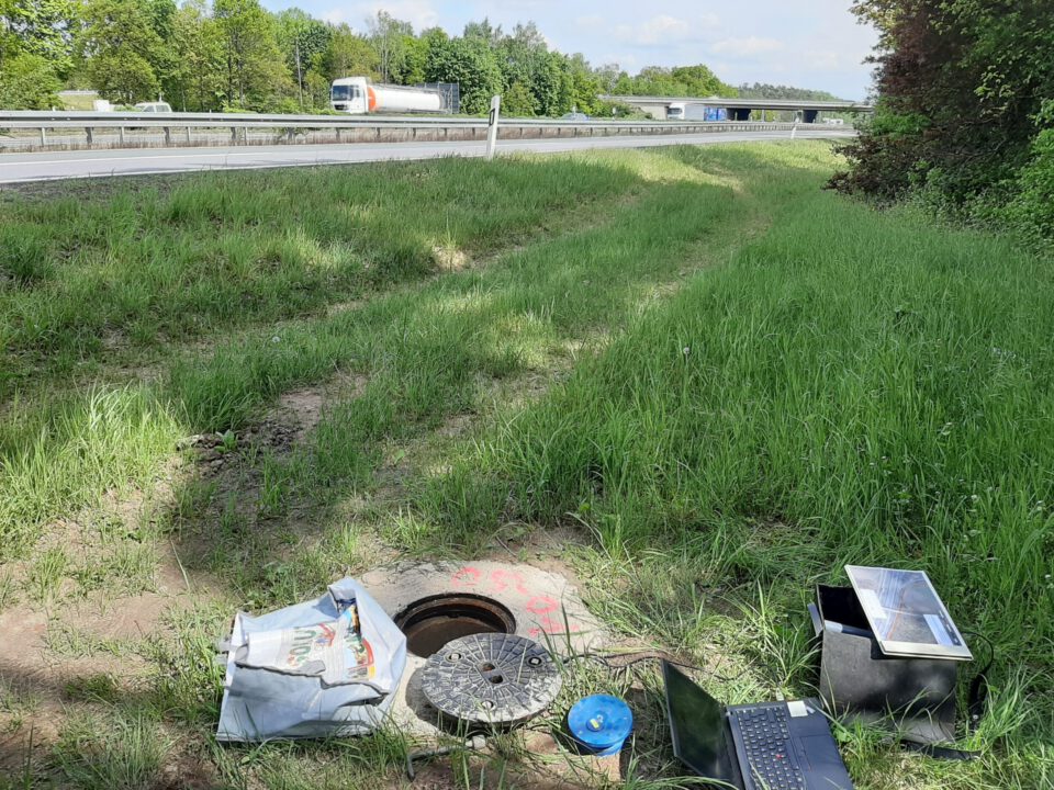 Das Bild zeigt eine grasbewachsene Fläche neben einer Autobahn, auf der Fahrzeuge vorbeifahren. Im Vordergrund befindet sich ein offener Kanalschacht mit dem Deckel daneben sowie verschiedene Gegenstände wie ein Laptop, ein blaues Messgerät und eine Tasche. Die Szene deutet auf eine Wartungs- oder Messaktivität im Außeneinsatz hin.