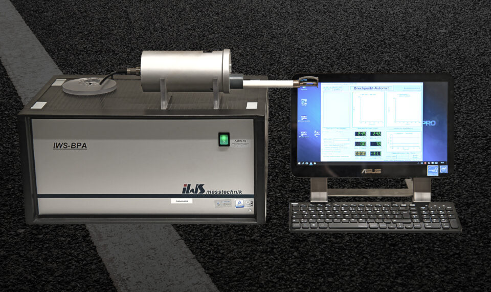 Das Bild zeigt ein Laboraufbau mit einem Messgerät vom Typ IWS-BPA, das mit einem ASUS-Computer verbunden ist. Auf dem Bildschirm sind verschiedene Diagramme und Daten zu sehen, die vermutlich Messungen oder Analysen darstellen. Der Aufbau befindet sich auf einer dunklen, strukturierten Oberfläche, im Hintergrund ist eine weiße Linie erkennbar.