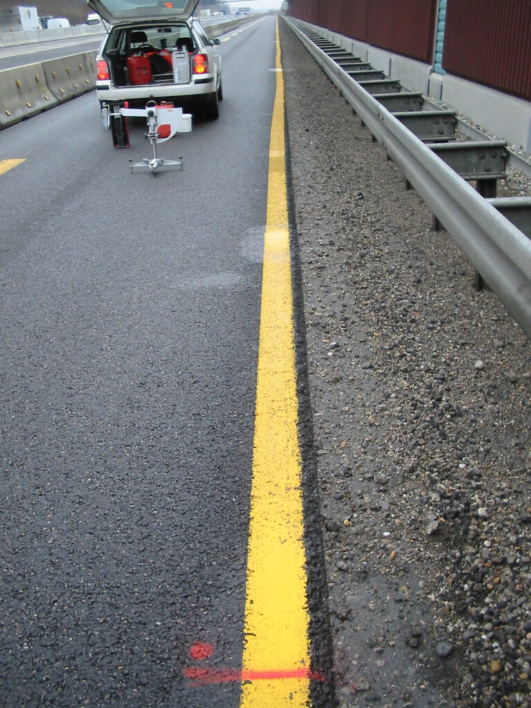 Das Bild zeigt einen Abschnitt einer Autobahn mit einer gelben Linie am Fahrbahnrand. Auf der linken Seite steht ein Auto mit geöffnetem Kofferraum, an dem ein Gerät angebracht ist, vermutlich für Wartungs- oder Prüfzwecke. Rechts neben der Straße befinden sich ein Leitplanke und ein geschotterter Seitenstreifen.