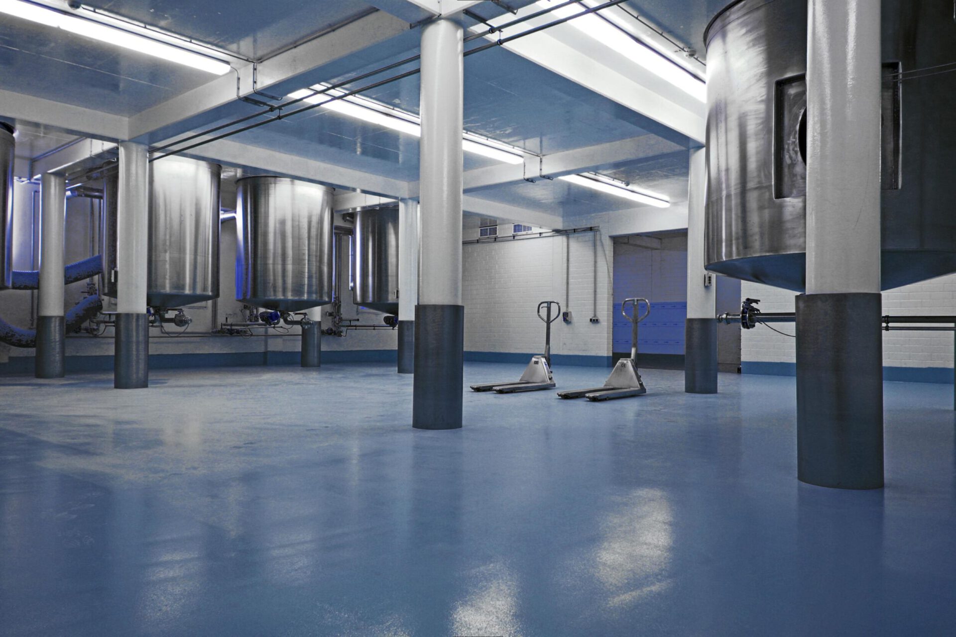 Das Bild zeigt eine industrielle Anlage mit großen Edelstahltanks, vermutlich zur Verarbeitung oder Lagerung von Flüssigkeiten. Der Boden ist mit einem blauen Belag beschichtet, und mehrere weiße Säulen stützen die Decke. Auf der rechten Seite sind zwei Hubwagen zu sehen.