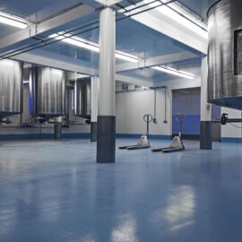 Das Bild zeigt eine industrielle Anlage mit großen Edelstahltanks, vermutlich zur Verarbeitung oder Lagerung von Flüssigkeiten. Der Boden ist mit einem blauen Belag beschichtet, und mehrere weiße Säulen stützen die Decke. Auf der rechten Seite sind zwei Hubwagen zu sehen.