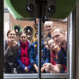 Das Bild zeigt eine Gruppe von Personen, die durch eine Glasscheibe eine Maschine bei einer Bohr- oder Schneidoperation beobachten. Im Zentrum ist ein Bohrkopf zu erkennen, der auf ein Werkstück gerichtet ist. Die Personen stehen dicht beieinander.