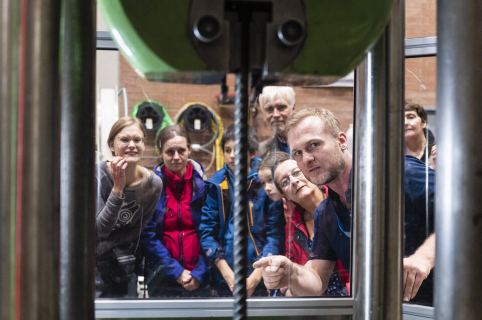Das Bild zeigt eine Gruppe von Personen, die durch eine Glasscheibe eine Maschine bei einer Bohr- oder Schneidoperation beobachten. Im Zentrum ist ein Bohrkopf zu erkennen, der auf ein Werkstück gerichtet ist. Die Personen stehen dicht beieinander.