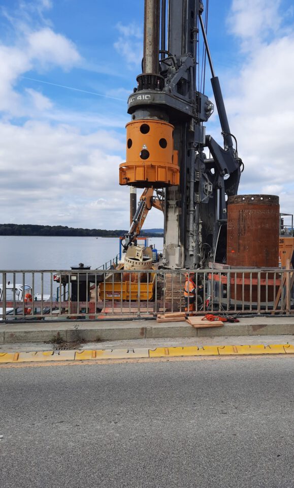 Das Bild zeigt eine Baustelle an einem Gewässer mit einem großen Bohrgerät, das auf einer Plattform am Ufer steht. Mehrere Arbeiter mit Sicherheitskleidung sind zu sehen, im Hintergrund erstreckt sich das Wasser mit einer Landzunge am Horizont.