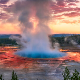 Das Bild zeigt eine geothermische heiße Quelle, aus deren Mitte Dampf aufsteigt. Das Wasser ist in der Mitte leuchtend blau und von rot-orangenen Farbtönen am Rand umgeben. Im Hintergrund sind Bäume, Hügel und ein farbenfroher Himmel bei Sonnenauf- oder -untergang zu sehen.