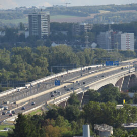 Das Bild zeigt eine Autobahnbrücke mit mehreren Fahrspuren, auf denen Autos und Lastwagen unterwegs sind. Die Brücke verläuft über eine grüne Landschaft mit Bäumen und Vegetation. Im Hintergrund sind hohe Gebäude, Wohnhäuser sowie hügelige Felder zu sehen.