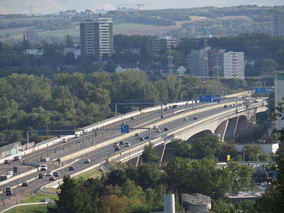 Das Bild zeigt eine Autobahnbrücke mit mehreren Fahrspuren, auf denen Autos und Lastwagen unterwegs sind. Die Brücke verläuft über eine grüne Landschaft mit Bäumen und Vegetation. Im Hintergrund sind hohe Gebäude, Wohnhäuser sowie hügelige Felder zu sehen.