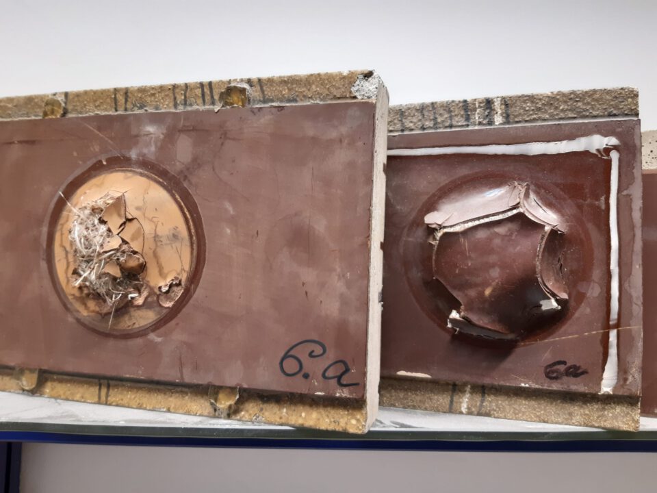 Das Bild zeigt zwei rechteckige Prüfkörper mit kreisförmigen Vertiefungen in der Mitte, die beschädigt wirken. Der linke Körper enthält in der Vertiefung Fasern und Rückstände, während der rechte Risse und Bruchstellen aufweist. Beide sind mit der Kennzeichnung 6.a beschriftet.