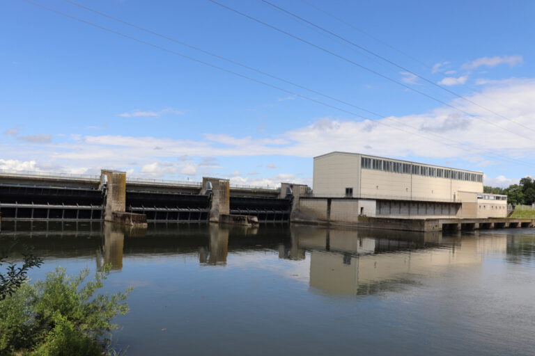 Das Bild zeigt ein Stauwerk mit mehreren Schleusen, das sich über ein Gewässer erstreckt. Rechts befindet sich ein großes Gebäude, das vermutlich zur technischen Infrastruktur des Damms gehört. Im Vordergrund ist etwas Vegetation zu sehen, darüber verlaufen Stromleitungen.